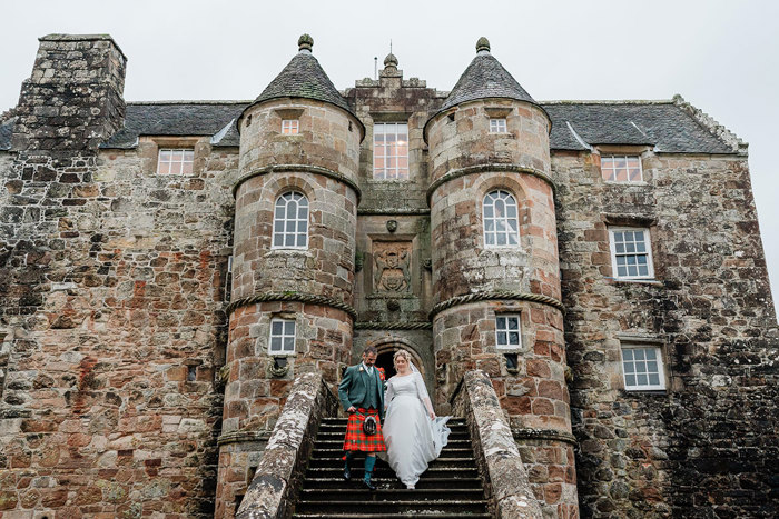 bride in white wedding dress and veil and groom in red and green tartan kilt outfit walk down outdoor castle staircase hand in hand