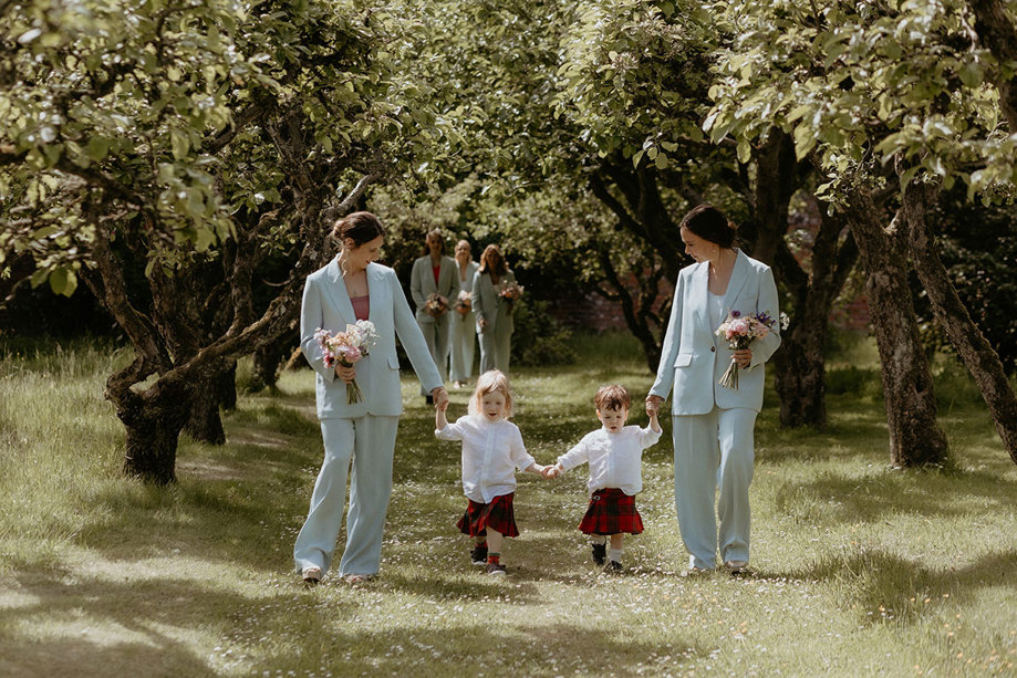 two bridesmaids wearing blue Whistles trouser suits walking down a natural outdoor aisle of trees with two toddlers wearing kilts during walled garden wedding ceremony at the Byre at Inchrya