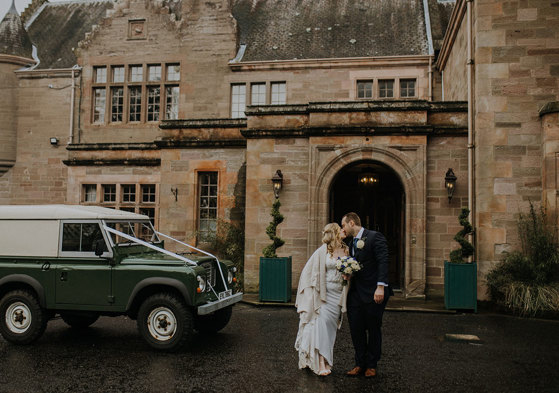 A bride and groom stand in front of a country house entrance and kiss with a green jeep next to them