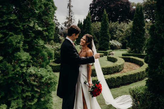 a smiling bride holding a bouquet of red roses with a groom in a lush green garden setting with manicured hedges at Achnagairn Castle