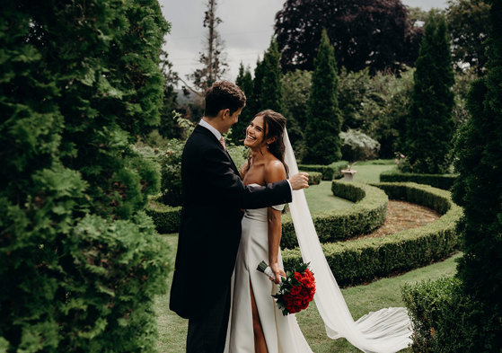 a smiling bride holding a bouquet of red roses with a groom in a lush green garden setting with manicured hedges at Achnagairn Castle