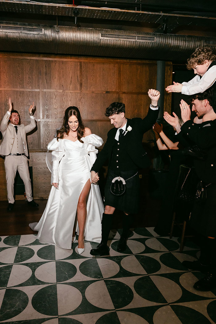 brunette bride in high leg slit wedding dress with puff sleeves and groom in full dark tartan kilt outfit walk holding hands out onto a black and white tiled dancefloor