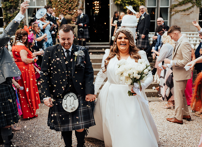 A man in a dark kilt holding hands with a bride in a long sleeved wedding dress walking past their guests who throw confetti over them