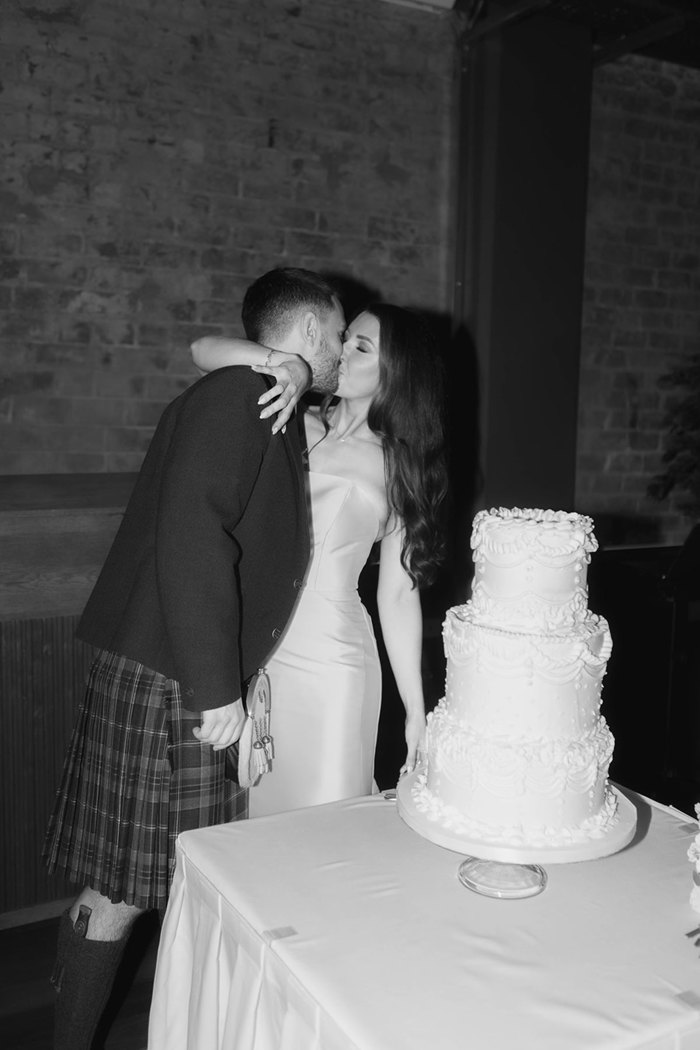 A bride and groom kiss standing next to their wedding cake