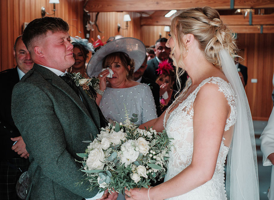 a groom cries as he greets a bride and holds her hands. A person in a large hat dabs a hanky to her eye as she looks on