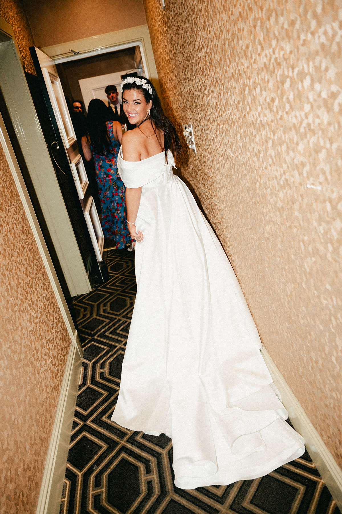 dark haired bride wearing floral headband and off the shoulder wedding dress looks back over her shoulder smiling in a carpeted hallway