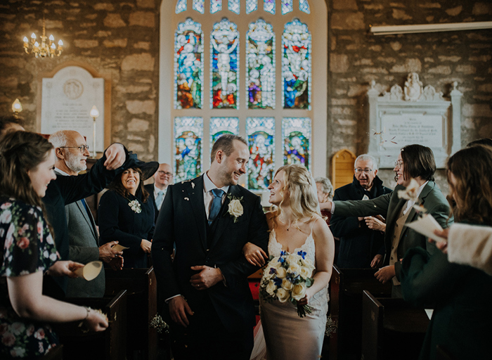 A bride and groom walking arm in arm out of a church looking at each other as confetti is being thrown on them