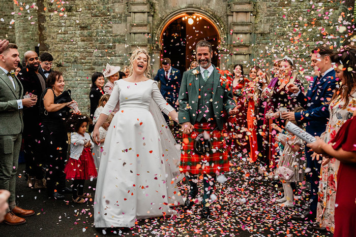 bride in white wedding dress and veil and groom in red and green tartan kilt step out of arched building opening hand in hand as pink heart shaped confetti is thrown on them