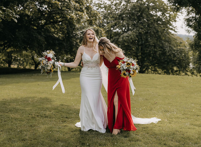 A bride in a white dress and a bridesmaid in a red dress stand with their arms around each other holding their bouquets in the other hand outside