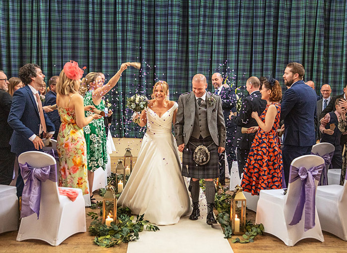 Wedding guests throw purple confetti over a bride and groom as they walk down a white aisle