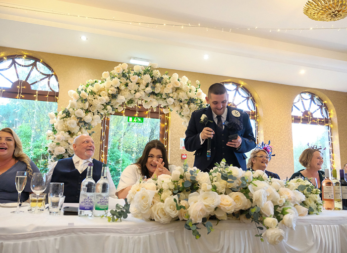 A groom making a speech holding a microphone and a phone while the bride and other people sitting at the table laugh