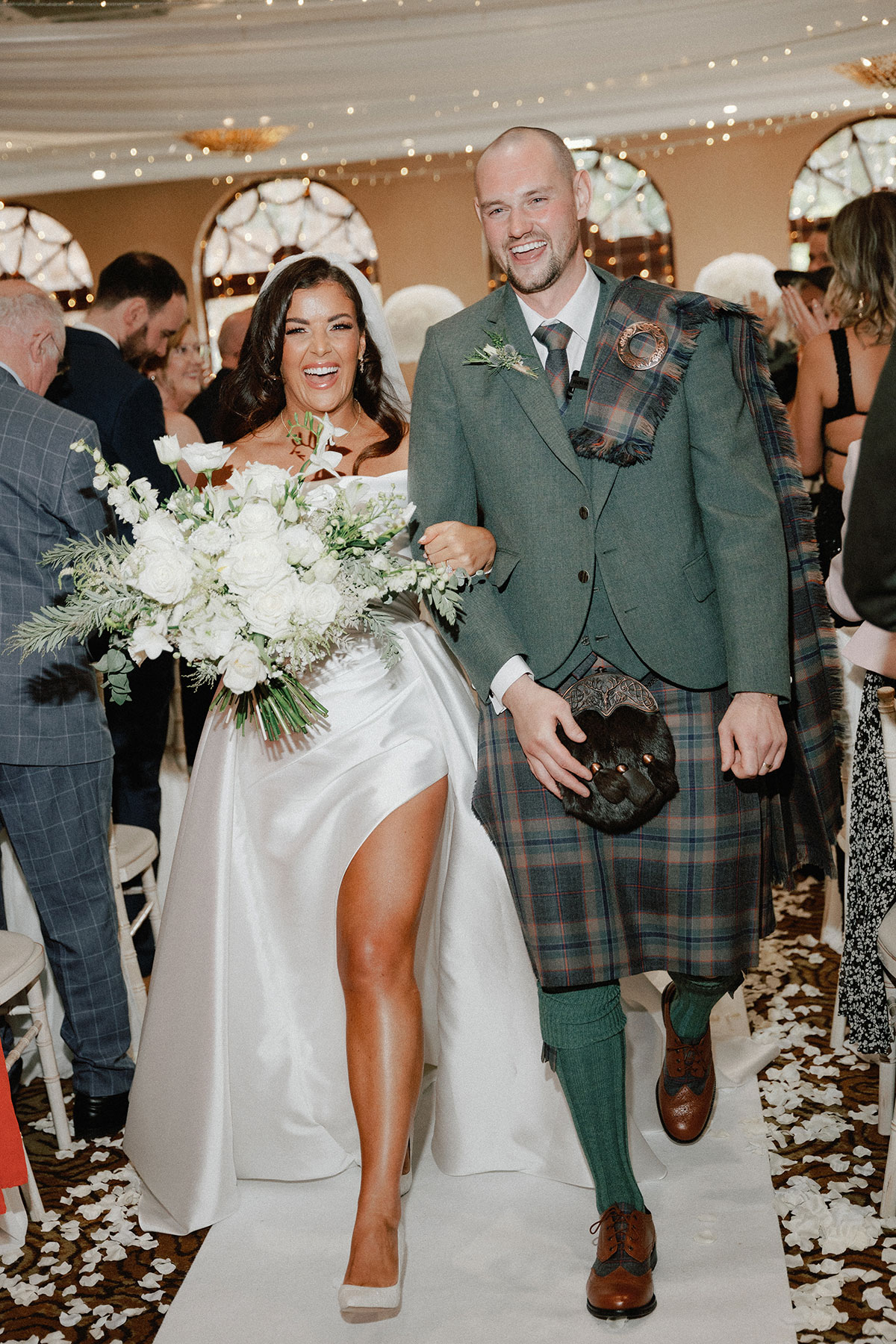 bride with veil and off the shoulder wedding dress with a leg slit stands holds a large white bouquet in one hand, holding onto groom in green, blue and purple tartan kilt outfit with green jacket as they walk up the aisle, confetti on the floor all around them