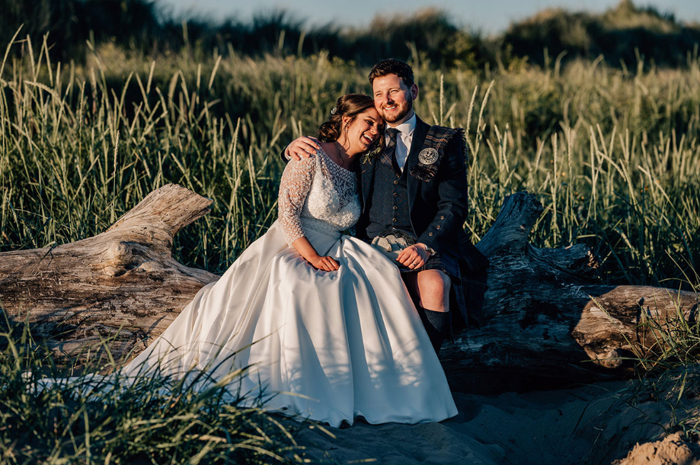 Bride and groom sit on log on beach and hug 