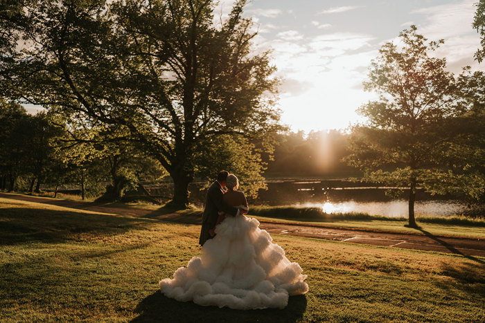 A bride wearing a wedding dress with a voluminous tulle skirt standing next to a groom wearing a green kilt look at the sun setting over trees and a pond