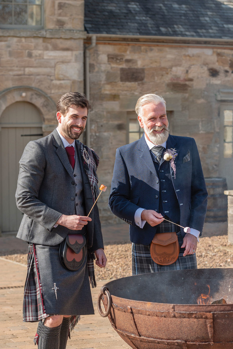 two men wearing kilt outfits from Eight Yards toasting marshmallows over the fire pit at Rosebery Steading