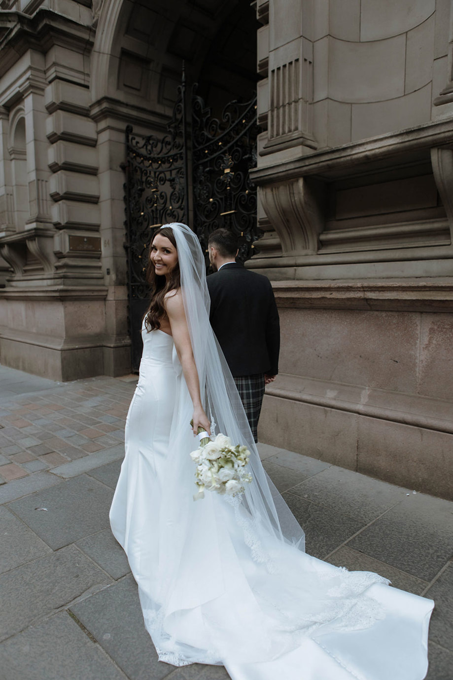 A bride looking over her shoulder at the camera as she walks down a street holding hands with the groom