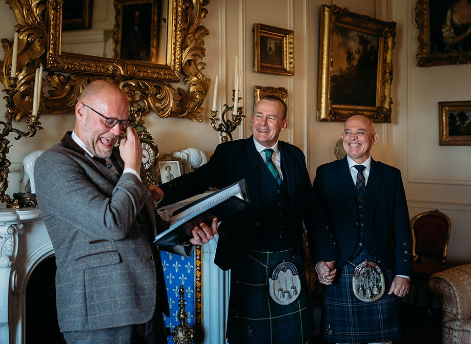 two grooms holding hands during a wedding ceremony as emotional celebrant laughs and wipes away a tear. There is a fireplace and pictures with gold frames in the background