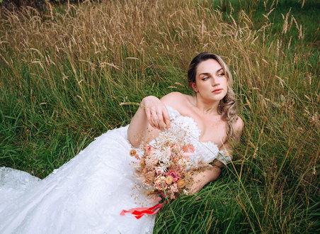 bride in off the shoulder dress lies in a field holding a pink and neutral coloured dried bouquet