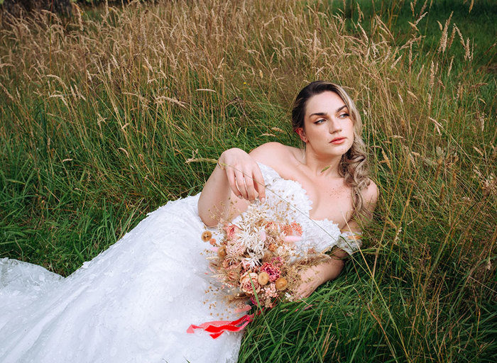 bride in off the shoulder dress lies in a field holding a pink and neutral coloured dried bouquet