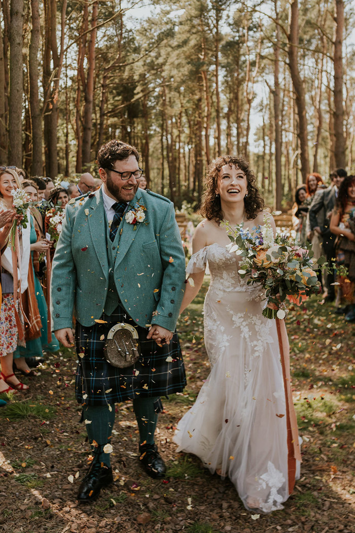 bride in off the shoulder flowy floral gown and groom in teal jacket and dark blue tartan kilt laugh and hold hands while looking up at the dried leaf confetti that falls around them