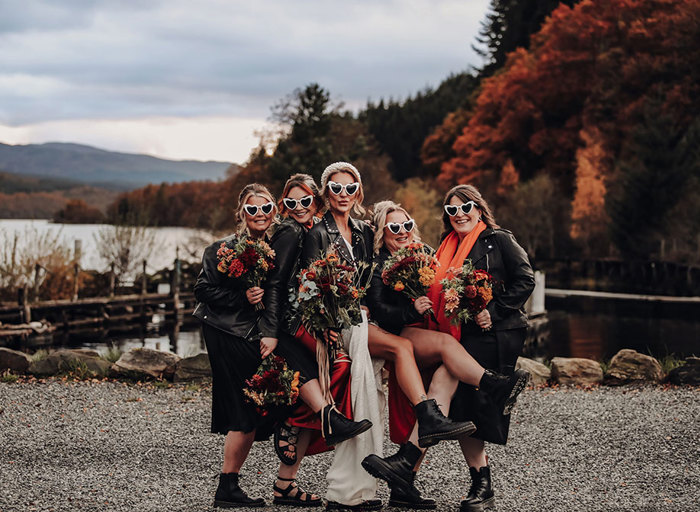 a bride and bridesmaids posing wearing heart shaped sunglasses, black leather jackets and black leather boots with autumn trees and landscape in background