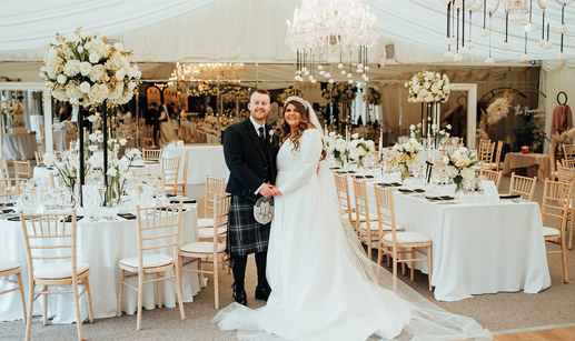 A man in a dark kilt holding hands with a bride in a long sleeved wedding dress as they stand inside a marquee with lots of tables, chairs, white flowers and chandeliers 