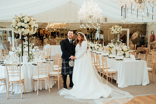 A man in a dark kilt holding hands with a bride in a long sleeved wedding dress as they stand inside a marquee with lots of tables, chairs, white flowers and chandeliers 