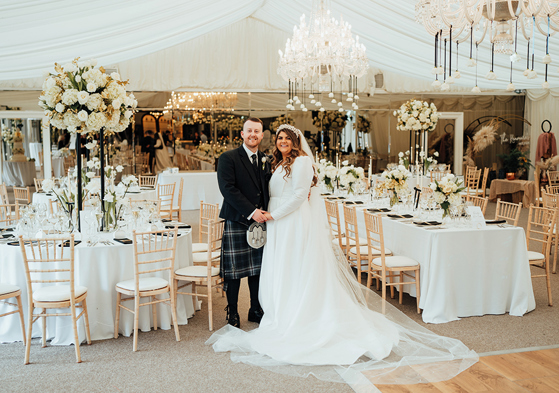 A man in a dark kilt holding hands with a bride in a long sleeved wedding dress as they stand inside a marquee with lots of tables, chairs, white flowers and chandeliers 