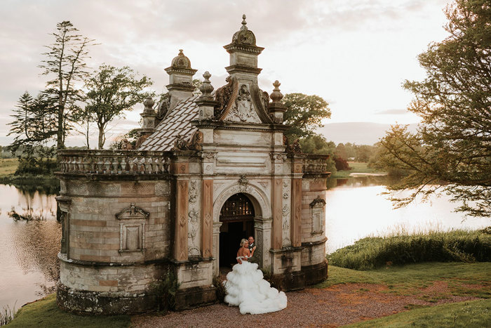 A bride wearing a wedding dress with a voluminous tulle skirt standing next to a groom wearing green stand in the door way of a small building in front of a large pond