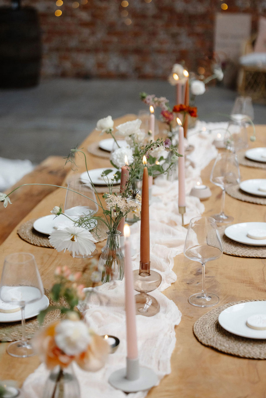 a long wooden dining table is decorated with a white linen runner, pale pink and burnt orange candlesticks, bud vases, rattan placemats and tableware