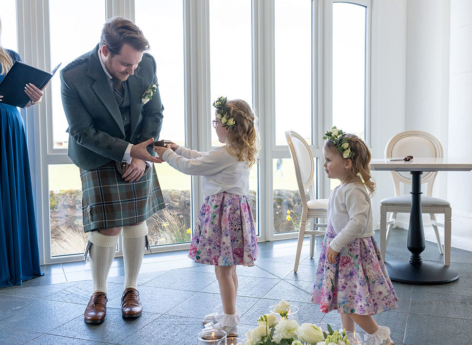 a girl wearing a pink flower dress and white cardigan passes a box to a man in a kilt as another young girl wearing a pink flower dress and white cardigan also watches on