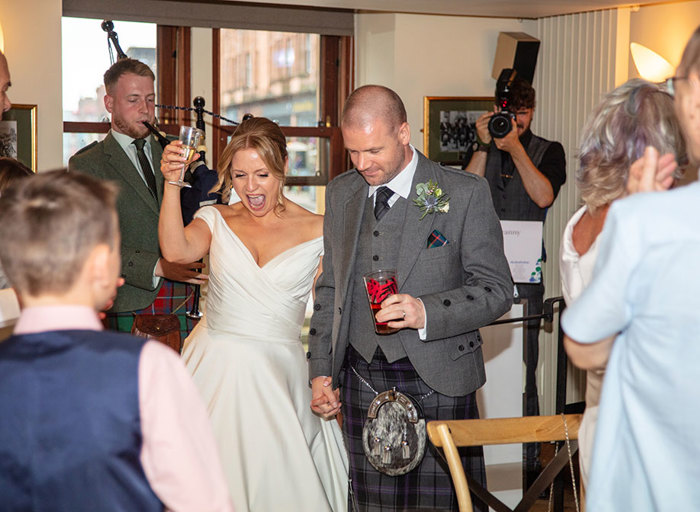 A bride and groom hold hands walking through a crowd holding glasses of drinks with a photographer and a bagpiper in the background