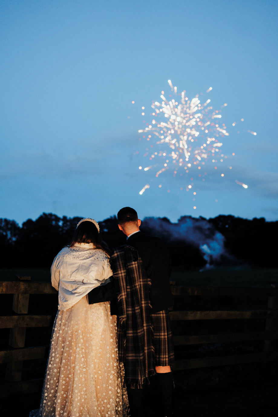 A man in a kilt and a woman in a white dress and jacket face away from the camera looking at a firework in the night sky