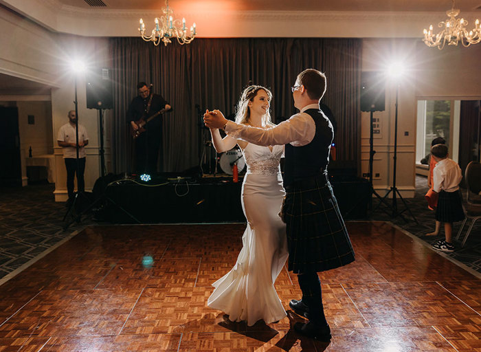 A bride in a wedding dress and a groom in a kilt dance together on a wooden dance floor in front of a small stage with a band