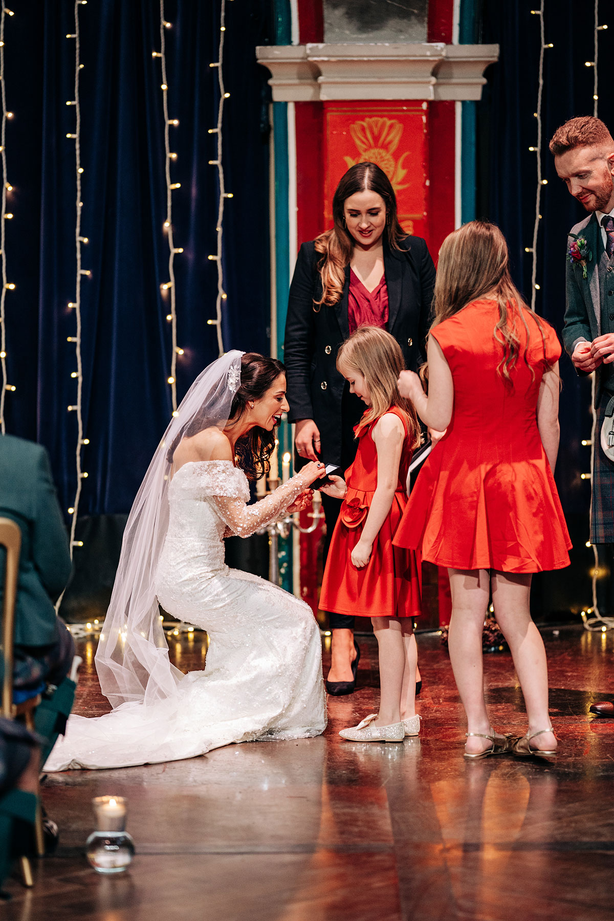 bride bends down to hug flower girls during wedding ceremony at oran mor