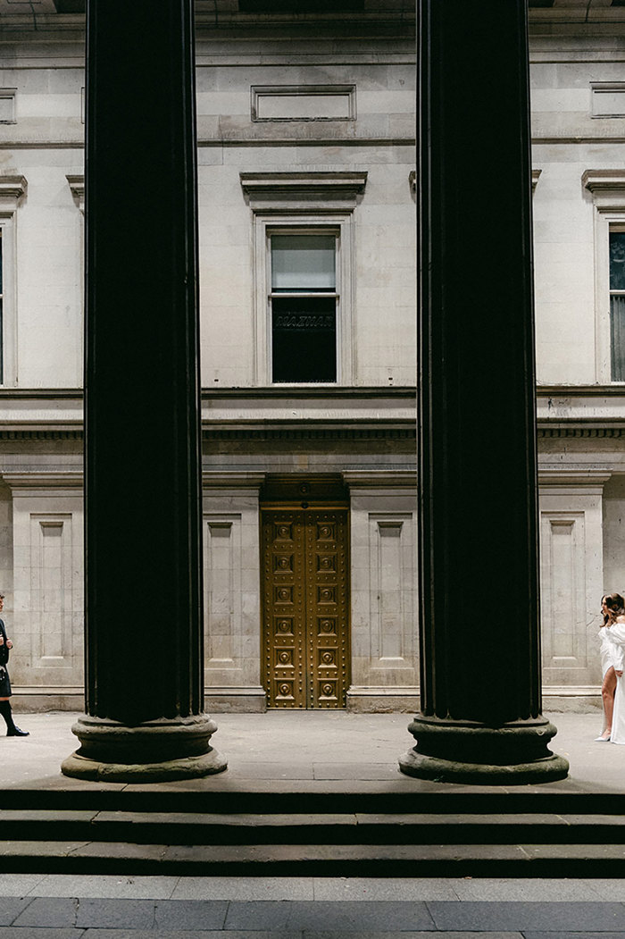 brunette bride in high leg slit wedding dress with puff sleeves and groom in full dark tartan kilt outfit walk from either sides of two large building columns