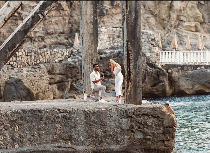 a man down on one knee proposing to a surprised woman on a rocky outcrop by the sea