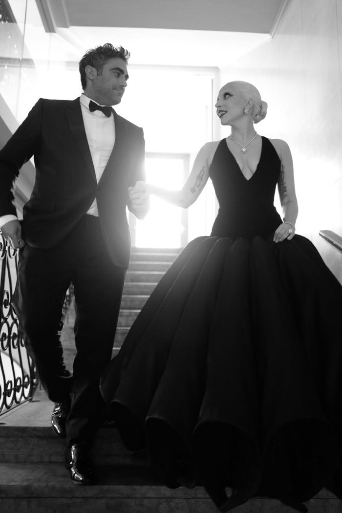 A black and white photo of Lady Gaga in a black ballgown and Michael Polansky in a tuxedo walking down stairs holding hands