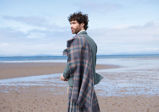 Man with long curled brown hair standing on beach wearing tartan plaid, blue jacket and kilt