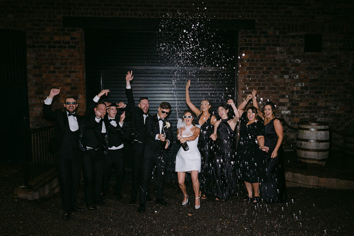 A group of people in black suits and black and white dresses. Two of them are spraying champagne from bottles.