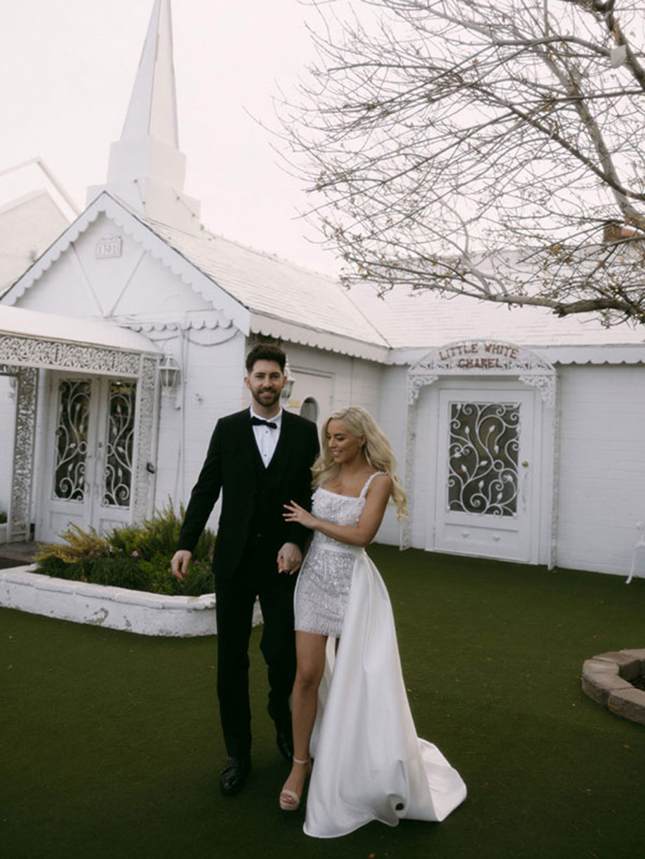 bride in a mini white dress with overskirt and groom in a classic tuxedo pose outside a little white wedding chapel