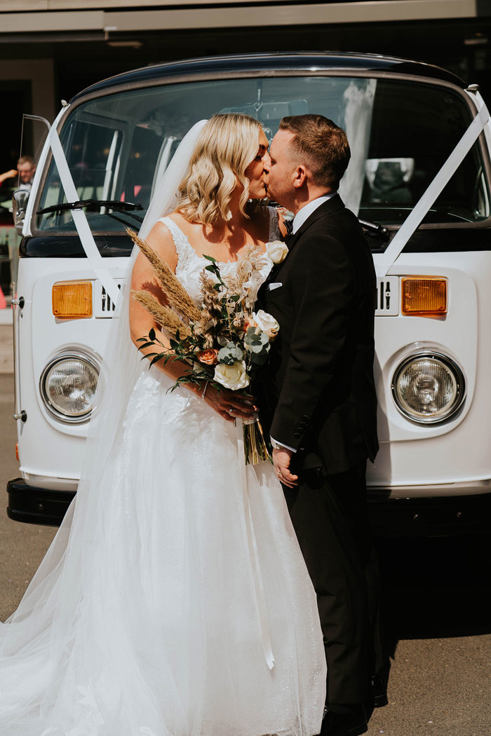 A bride and groom kissing in front of a white VW camper van.