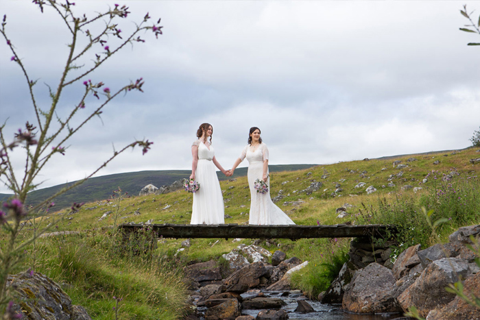 Two Brides Holding Hands And Standing On A Bridge In The Highlands