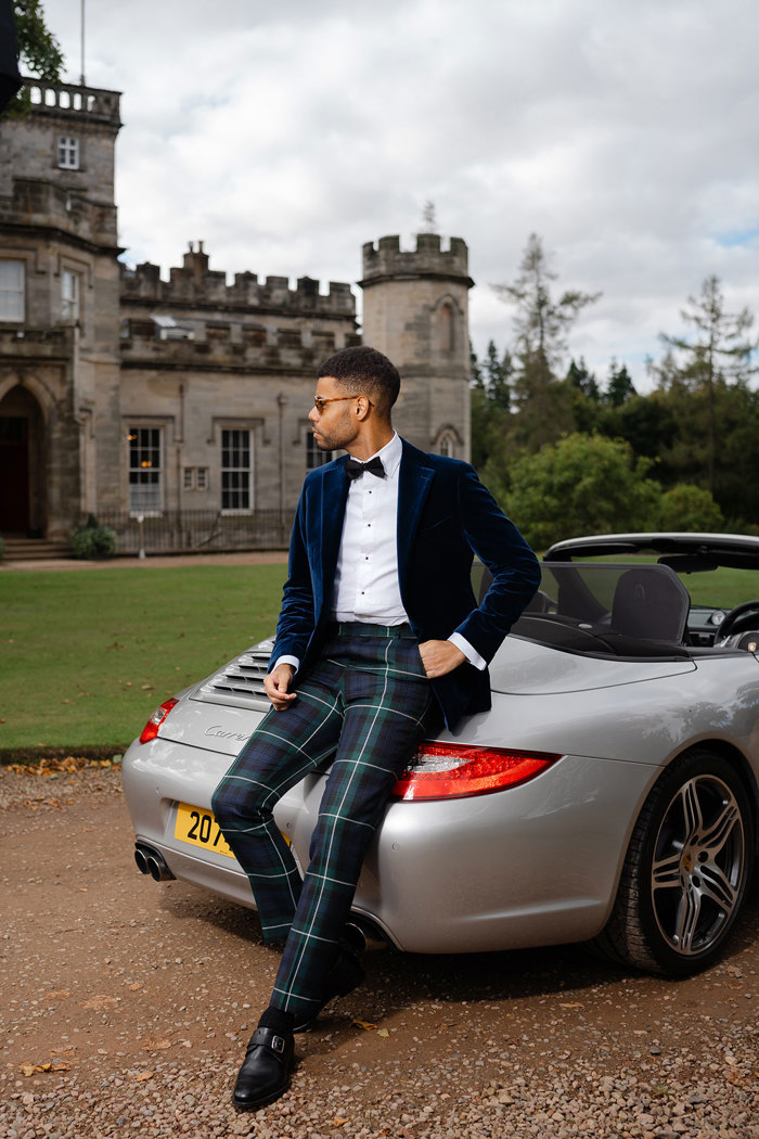 A man wearing green tartan trousers and a navy velvet suit jacket leans against a silver convertible car in front of a castle