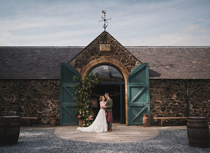 A bride and groom standing outside an arched doorway with green wooden doors at Byre at Inchyra