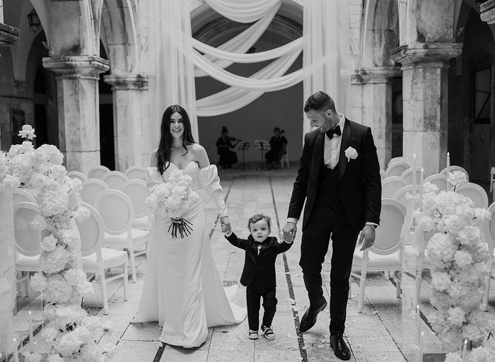 A couple in formal attire and young boy walking through Sponsa Palace Dubrovnik set for a wedding ceremony; the woman wears a white dress, the young boy a black suit, and the man dons a black tuxedo.