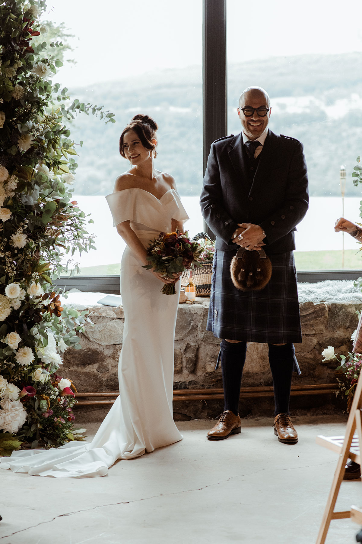 groom in blue, grey and white tartan kilt outfit stands in front of large scenic window with bride to his side in off the shoulder white wedding dress, holding a bouquet of flowers in front of her waist