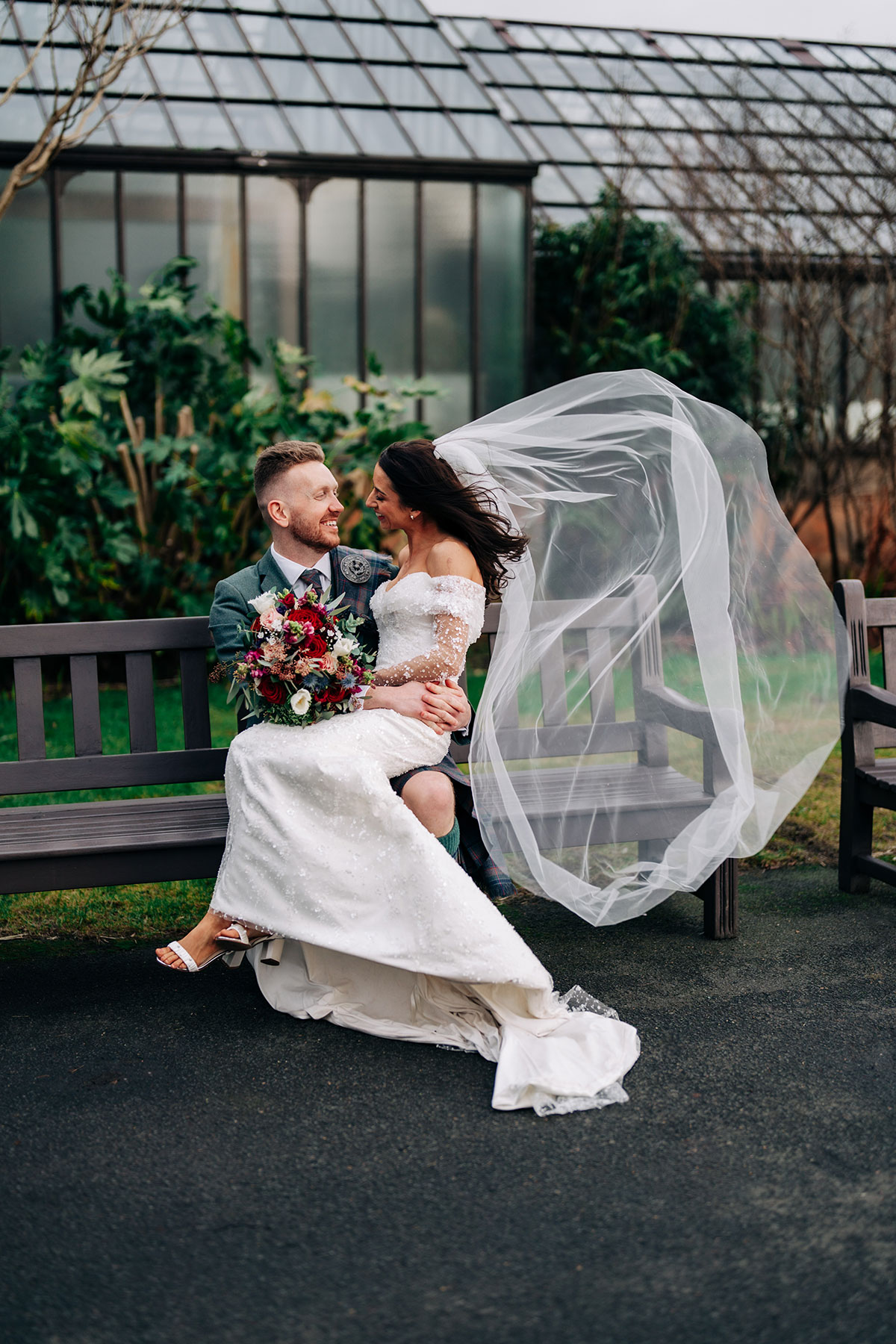 bride sits on groom's knee on bench at glasgow botanic gardens