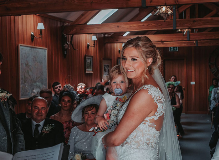 a bride holding a toddler in her arms during a wedding ceremony in a wooden clad room. Guests seated in rows watch on