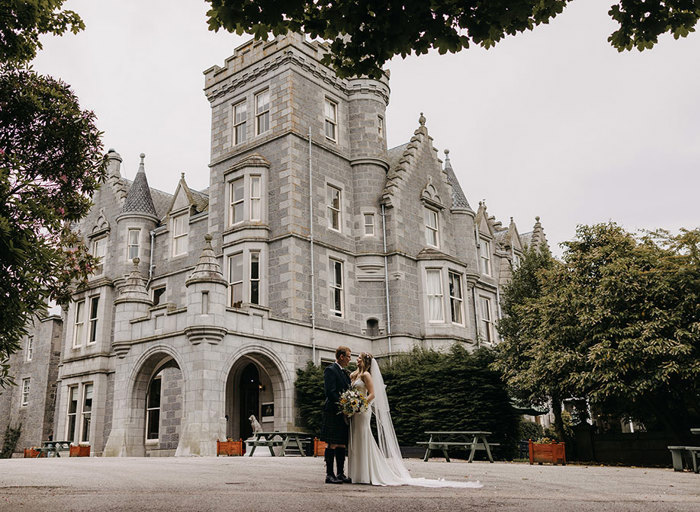 A bride and groom stand looking into each other's eyes outside a grand grey stone building